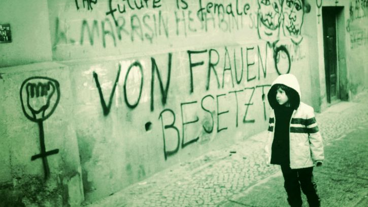 Film still from THE FUTURE IS FEMALE: A child stands in front of a house wall painted with numerous slogans and symbols. Among them are “The future is female” and “Occupied by women.” 