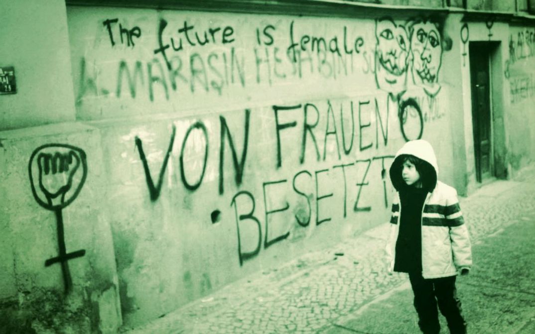 Film still from THE FUTURE IS FEMALE: A child stands in front of a house wall painted with numerous slogans and symbols. Among them are “The future is female” and “Occupied by women.” 