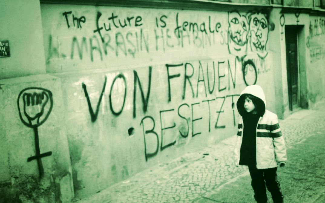 Filmstill aus THE FUTURE IS FEMALE: Ein Kind steht vor einer Hauswand, die mit zahlreichen Sprüchen und Symbolen bemalt ist. Darunter "The future is female" und "Von Frauen besetzt". 