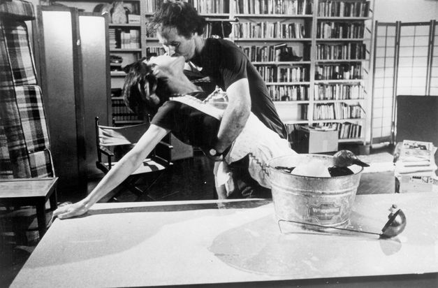 Film still from Yvonne Rainer’s „The Man Who Envied Women“. A women leans on a table, while she kisses a man who is holding her. On the table there is a wooden duck in a bucket and a ladle. In the room in the background there are bookshelves and armchairs.