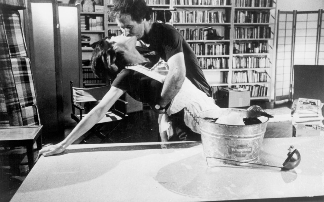 Film still from Yvonne Rainer’s „The Man Who Envied Women“. A women leans on a table, while she kisses a man who is holding her. On the table there is a wooden duck in a bucket and a ladle. In the room in the background there are bookshelves and armchairs.