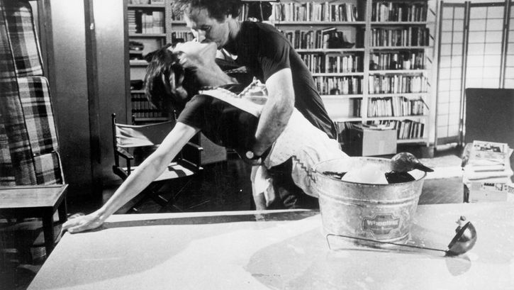 Filmstill aus dem Film „The Man Who Envied Women“ von Yvonne Rainer. Eine Frau stützt sich auf einem Tisch ab und küsst einen Mann, welcher sie im Arm hält. Auf dem Tisch liegt eine Holzente in einem Eimer und eine Suppenkelle. Im Hintergrund ein Zimmer mit Stühlen, Raumtrennern und einem Bücherregal.