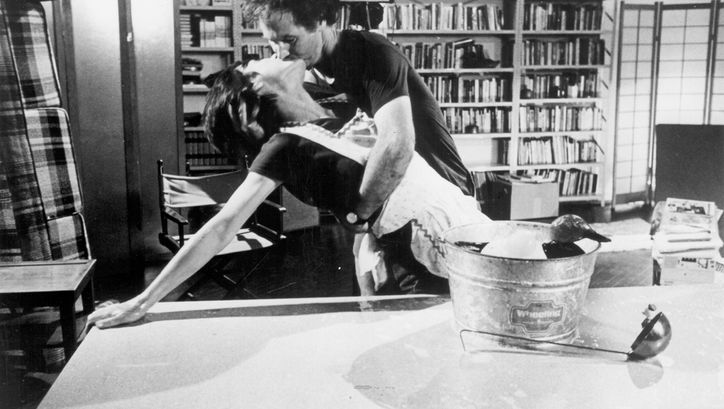 Filmstill aus dem Film „The Man Who Envied Women“ von Yvonne Rainer. Eine Frau stützt sich auf einem Tisch ab und küsst einen Mann, welcher sie im Arm hält. Auf dem Tisch liegt eine Holzente in einem Eimer und eine Suppenkelle. Im Hintergrund ein Zimmer mit Stühlen, Raumtrennern und einem Bücherregal.