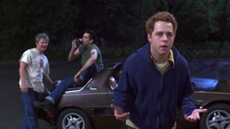 Film still from SUBURBIA: Three teenagers on the street, one of them sitting on a car.