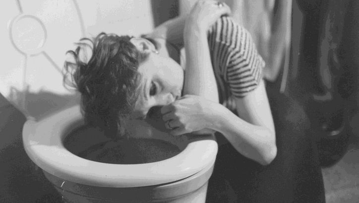 Film still from EIN GANZ UND GAR VERWAHRLOSTES MÄDCHEN: A woman with crooked hair crouches with her head above the toilet bowl. She looks dazed.