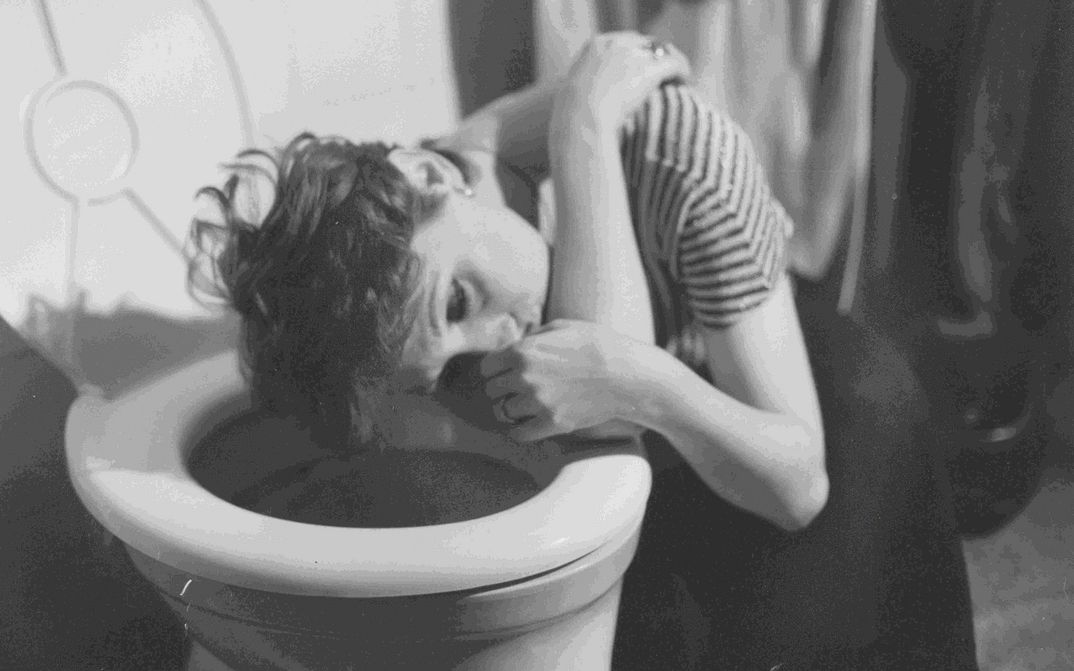 Film still from EIN GANZ UND GAR VERWAHRLOSTES MÄDCHEN: A woman with crooked hair crouches with her head above the toilet bowl. She looks dazed.