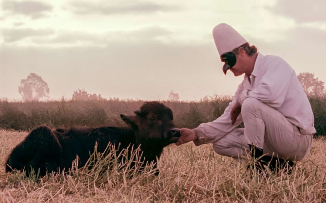 Filmstill aus BELLA E PERDUTA. Ein Mann mit einer Maske kniet vor einem liegenden Büffel.