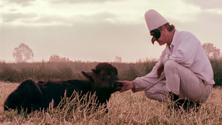Film still from BELLA E PERDUTA. A man with a mask kneels in front of a lying buffalo.
