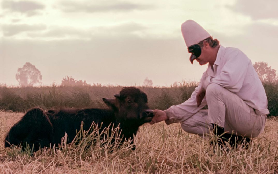 Film still from BELLA E PERDUTA. A man with a mask kneels in front of a lying buffalo.