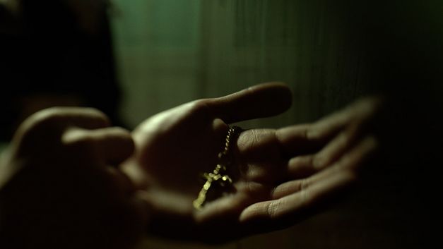 Filmstill from AFTER DREAMING by Christine Haroutounian. The picture shows two open hands holding a golden chain with a cross in the darkness.