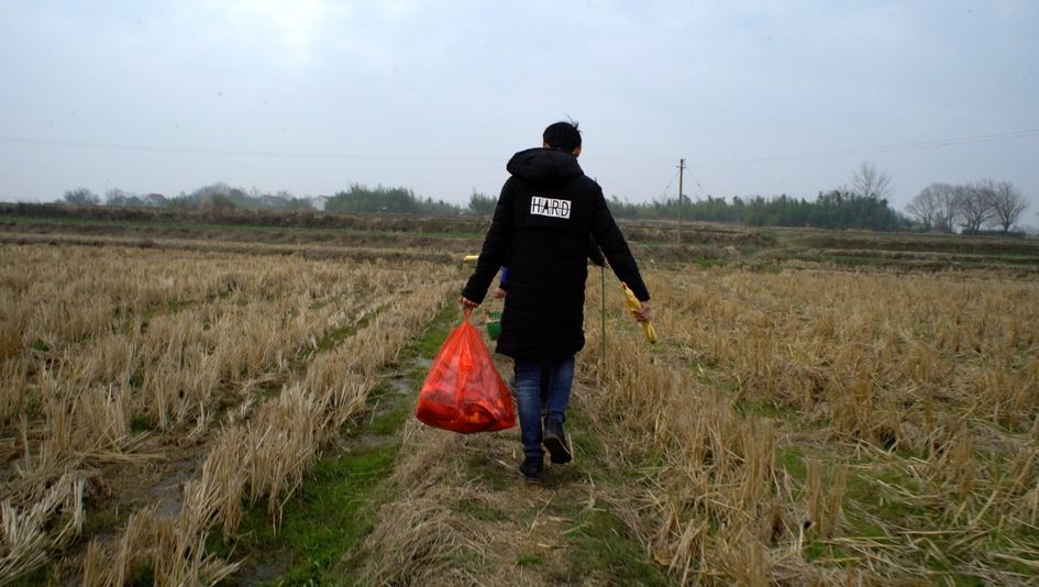 Filmstill aus YOUTH (HOMECOMING): Man sieht von hinten einen Menschen mit schwarzer Jacke und roter Plastiktüte über ein abgeerntetes Feld gehen. 