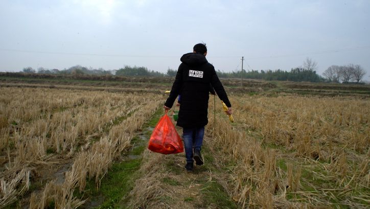 Filmstill aus YOUTH (HOMECOMING): Man sieht von hinten einen Menschen mit schwarzer Jacke und roter Plastiktüte über ein abgeerntetes Feld gehen. 