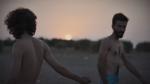 Zwei Männer mit nacktem Oberkörper gehen bei Sonnenuntergang über einen offenen Strand oder eine weite Ebene.