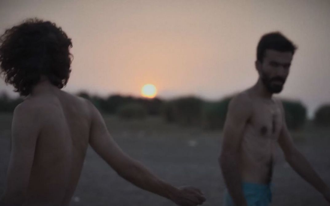 Zwei Männer mit nacktem Oberkörper gehen bei Sonnenuntergang über einen offenen Strand oder eine weite Ebene.