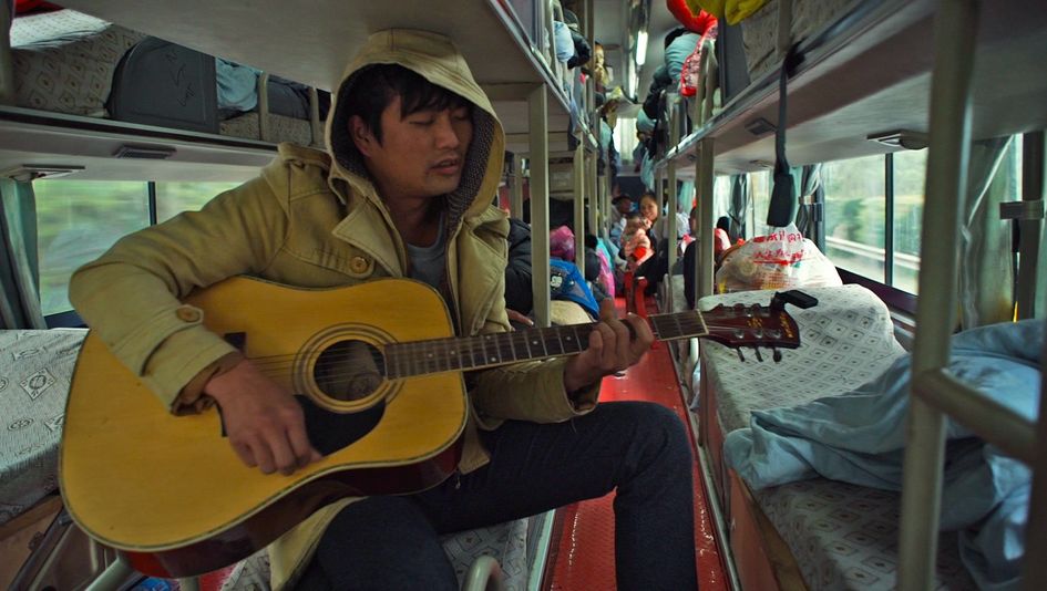 Filmstill aus YOUTH (HARD TIMES): Ein junger Mann mit einer Jacke mit Kapuze spielt in einem Nachtzug Gitarre. Er sitzt in einem Stockbett, im Hintergrund sind weitere Betten und Menschen zu sehen.
