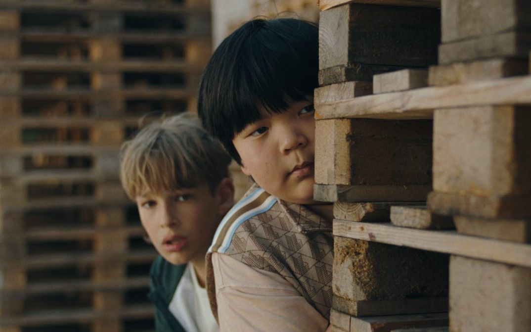 Film still from DER PRANK: Two boys peek out from behind a stack of pallets.