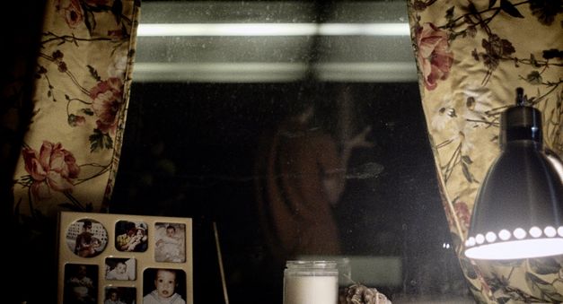 Still from the film "Happer’s Comet" by Tyler Taormina. A window sill at night: family photos in a frame, a candle, a lamp. In the background we see the reflection of a person in an orange shirt. 