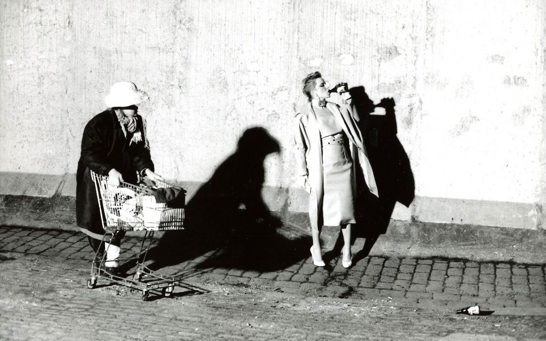 Film still from BILDNIS EINER TRINKERIN: An elegantly dressed woman drinks beer from a bottle on the street. Another woman with a fully loaded shopping cart walks by.