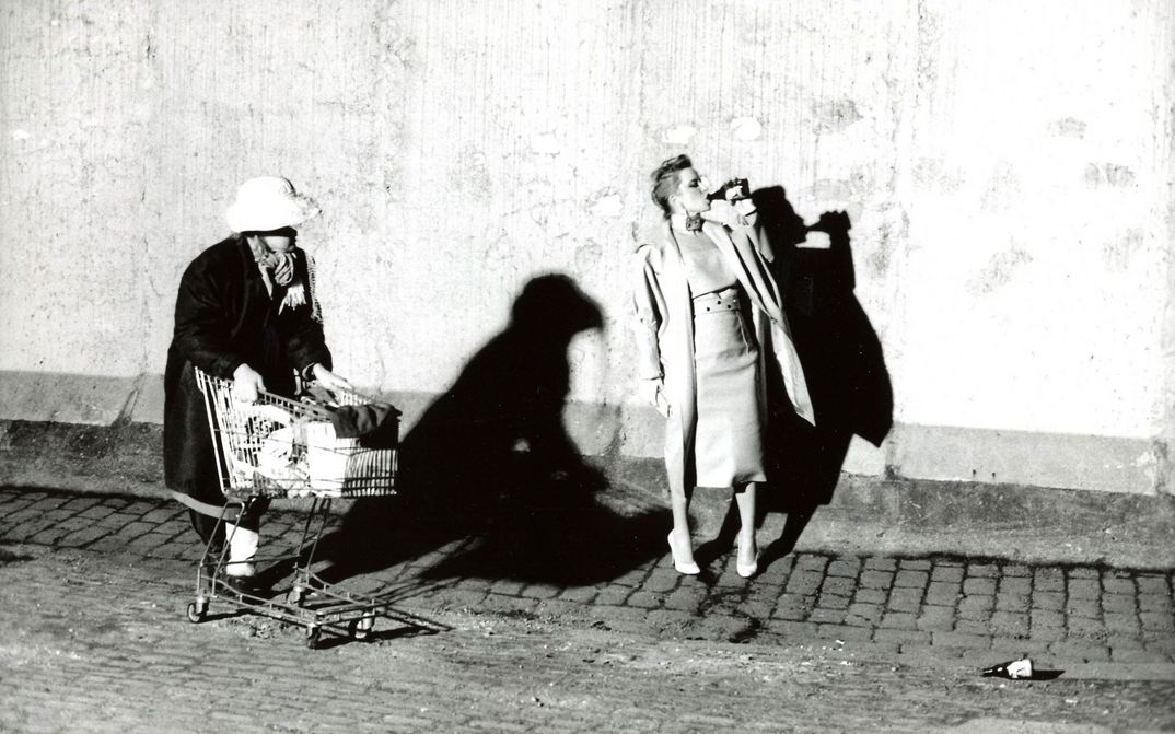 Filmstill aus BILDNIS EINER TRINKERIN: Eine elegant gekleidete Frau trinkt auf der Straße Bier aus einer Flasche. Eine andere Frau mit einem vollbepackten Einkaufswagen kommt daran vorbei.