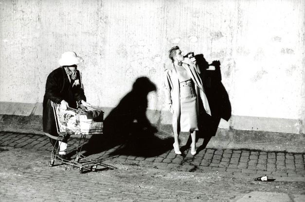 Filmstill aus BILDNIS EINER TRINKERIN: Eine elegant gekleidete Frau trinkt auf der Straße Bier aus einer Flasche. Eine andere Frau mit einem vollbepackten Einkaufswagen kommt daran vorbei.
