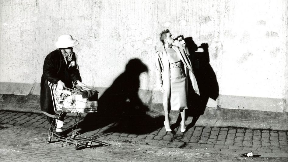 Film still from BILDNIS EINER TRINKERIN: An elegantly dressed woman drinks beer from a bottle on the street. Another woman with a fully loaded shopping cart walks by.