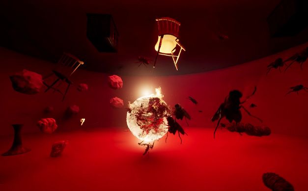 Film still from Assem Hendawi’s "Simia: Stratagem for Undestining". A round, low room in red light. In the center a glowing fireball, around it flying objects (stones, chairs), a hand and insects.