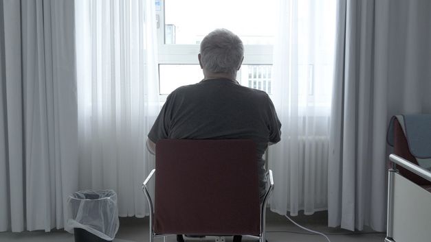 Film still from PALLIATIVSTATION (Palliative Care Unit) by Philipp Döring. The image shows a person in a hospital room on a chair in front of the window with their back to the camera.