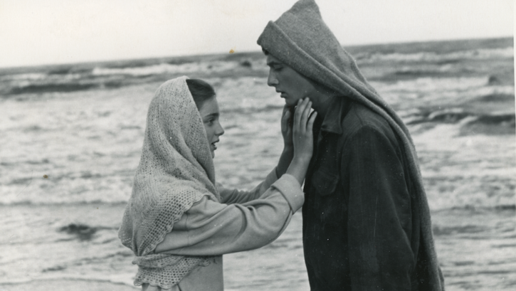 Film still fromm I SEE THE SUN: A girl and a boy stand facing each other in front of the sea, she reaches for his face.