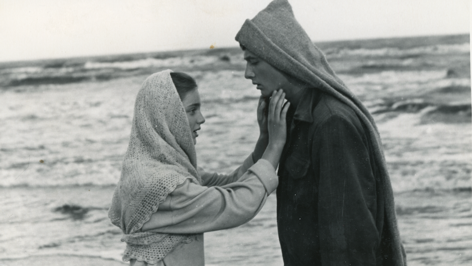 Filmstill aus I SEE THE SUN: Ein Mädchen und ein Junge stehen einander zugewandt vor dem Meer, sie greift nach seinem Gesicht.