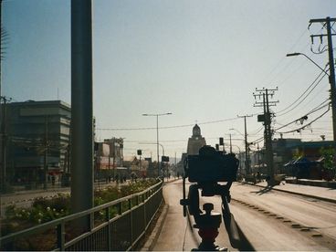 A city street. Emerging from the bottom of the frame is the silhouette of a camera on a tripod. In the centre of the frame, in the distance, is a church.