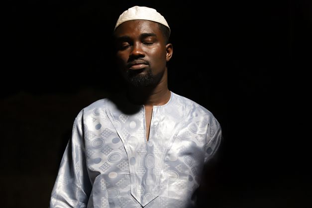 Film still from Eric Gyamfi’s film “CERTAIN WINDS FROM THE SOUTH”. A person stands in a pool of light against a black background with their eyes closed, wearing a kanzu and a kofia. 