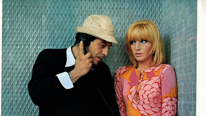 Film still from LE FATE: A man and a woman are standing in a blue-panelled telephone booth. He holds the telephone receiver in his hand. She is wearing a strikingly patterned dress in orange-pink. 