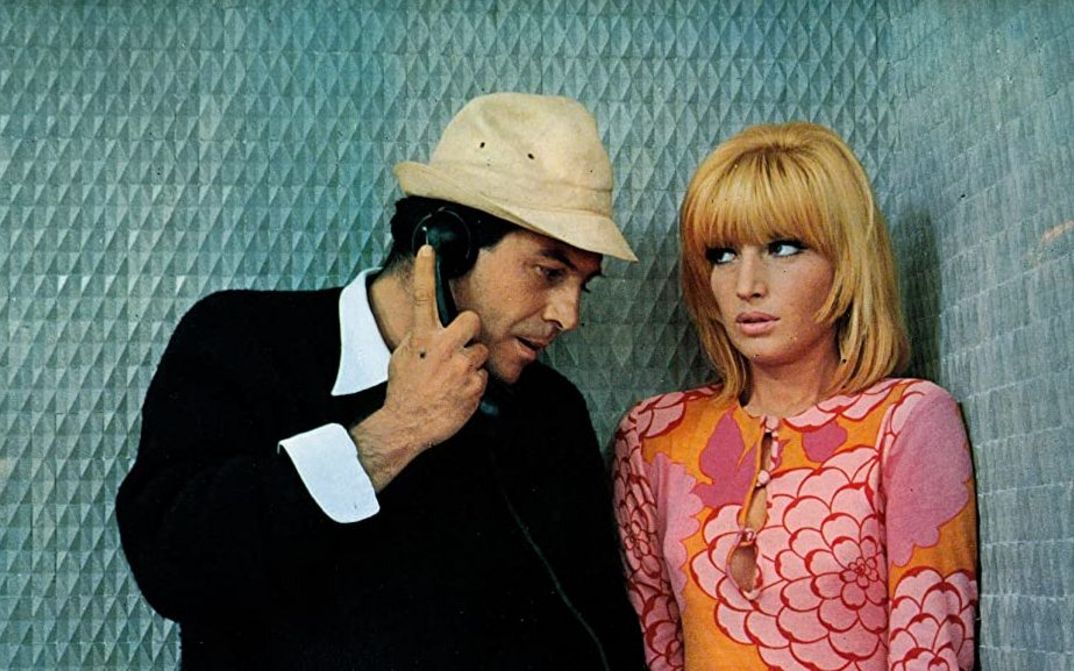 Film still from LE FATE: A man and a woman are standing in a blue-panelled telephone booth. He holds the telephone receiver in his hand. She is wearing a strikingly patterned dress in orange-pink.
