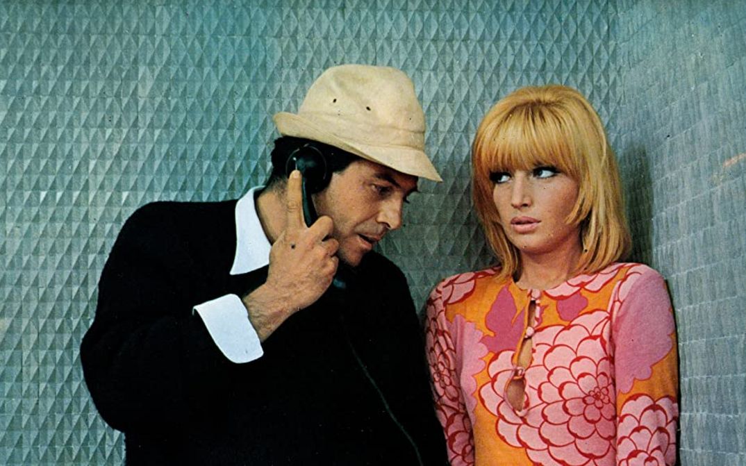 Film still from LE FATE: A man and a woman are standing in a blue-panelled telephone booth. He holds the telephone receiver in his hand. She is wearing a strikingly patterned dress in orange-pink. 