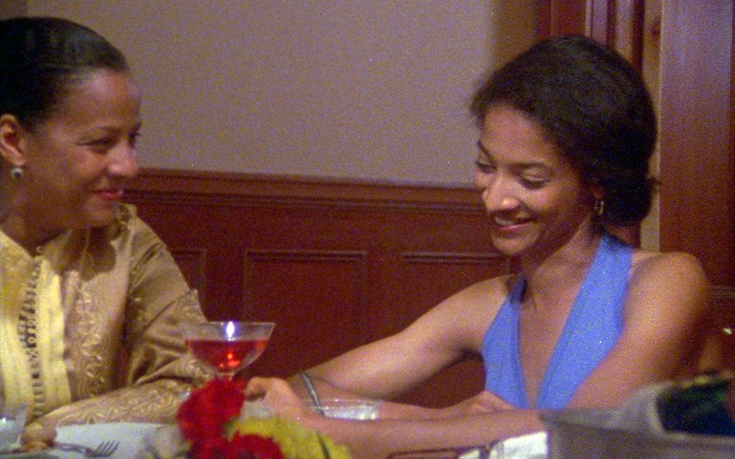 Film still from LOSING GROUND: Two women sit at a table laughing and chatting.