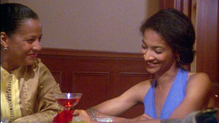 Film still from LOSING GROUND: Two women sit at a table laughing and chatting.