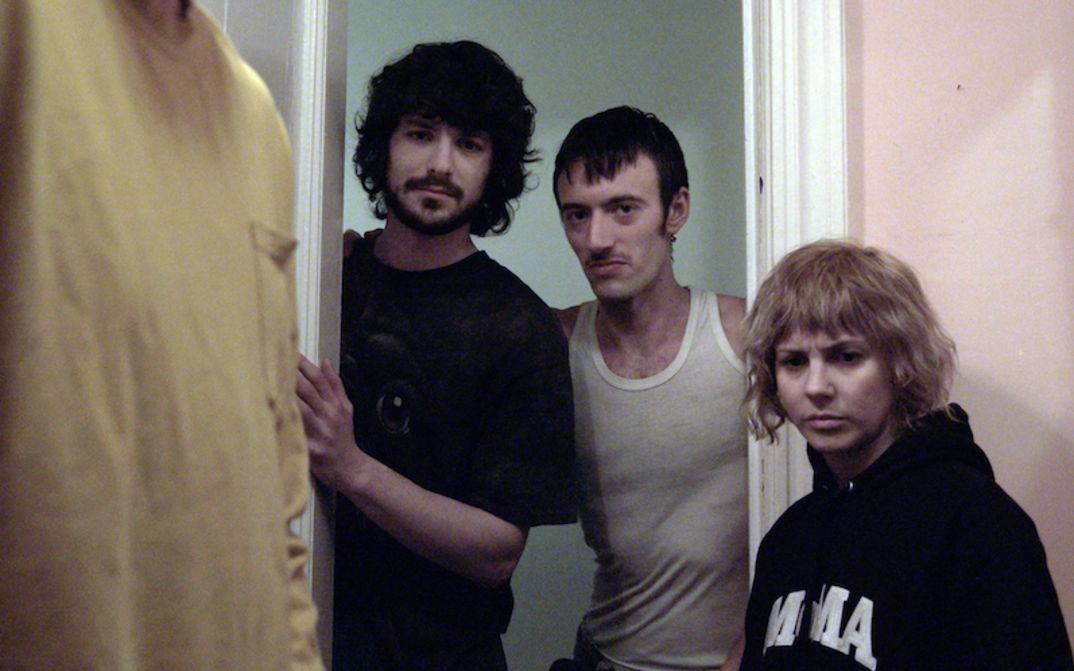 Film still from THE WRONG MOVIE: Three people are standing in a doorway looking into a room. Only part of another person can be seen there.