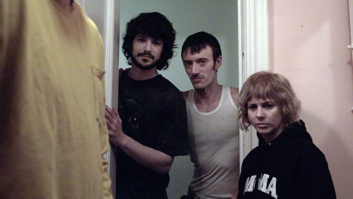 Film still from THE WRONG MOVIE: Three people are standing in a doorway looking into a room. Only part of another person can be seen there.
