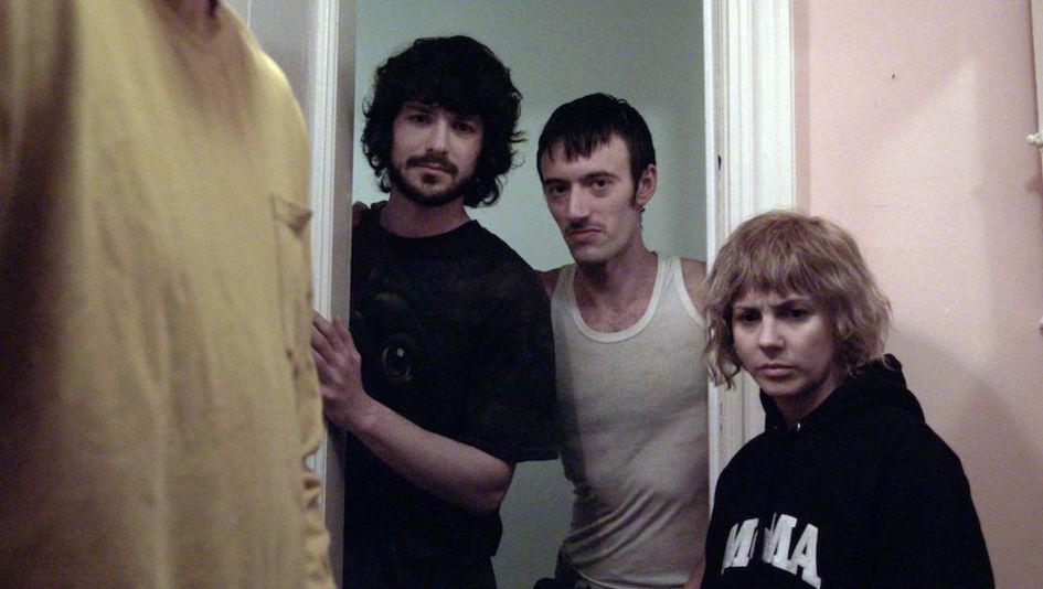 Filmstill aus THE WRONG MOVIE: Drei Personen stehen in einem Türrahmen und blicken in ein Zimmer. Dort sieht man nur angeschnitten eine weitere Person.