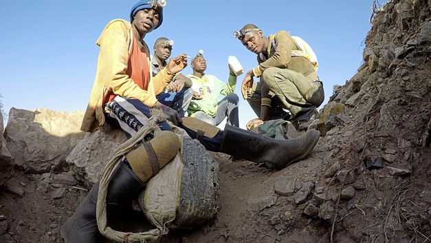 Still from the film "The Zama Zama Project" by Rosalind Morris. People with headlamps sit on the dirt floor.