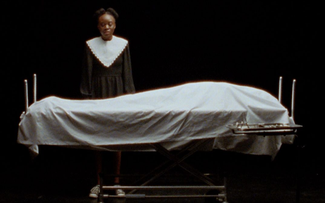 Still from the film "Cette Maison (This House)" by Miryam Charles. A teenage girl dressed in black and white stands behind a body covered by a sheet on a medical bed, in a black soundstage.