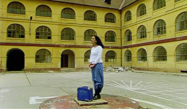 Filmstill aus "Diese Tage in Terezín" von Sibylle Schönemann. Zu sehen ist eine Frau in einem Innenhof auf einer Plattform mit einem Koffer. 