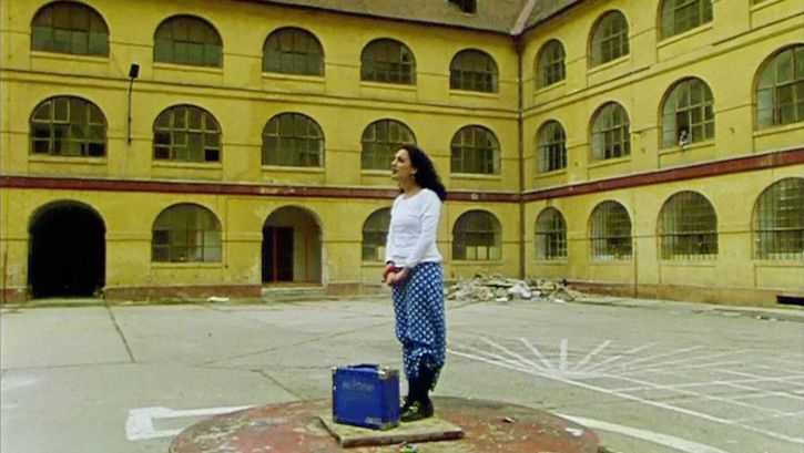 Film still from "Diese Tage in Terezín" by Sibylle Schönemann. It shows a woman in a courtyard on a platform with a suitcase.