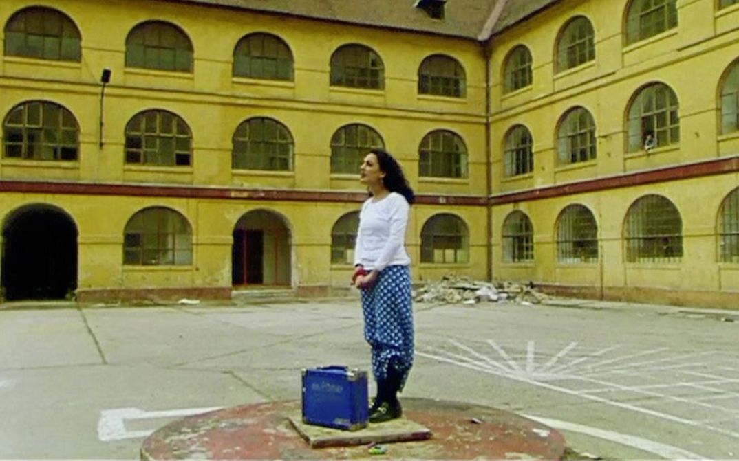 Film still from "Diese Tage in Terezín" by Sibylle Schönemann. It shows a woman in a courtyard on a platform with a suitcase.