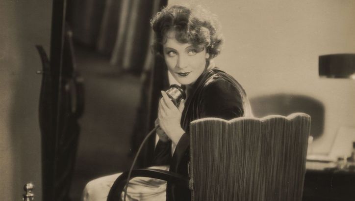 Film still from DIE FRAU, NACH DER MAN SICH SEHNT. Marlene Dietrich is sitting on a chair in elegant clothes and holding a telephone in her hand.