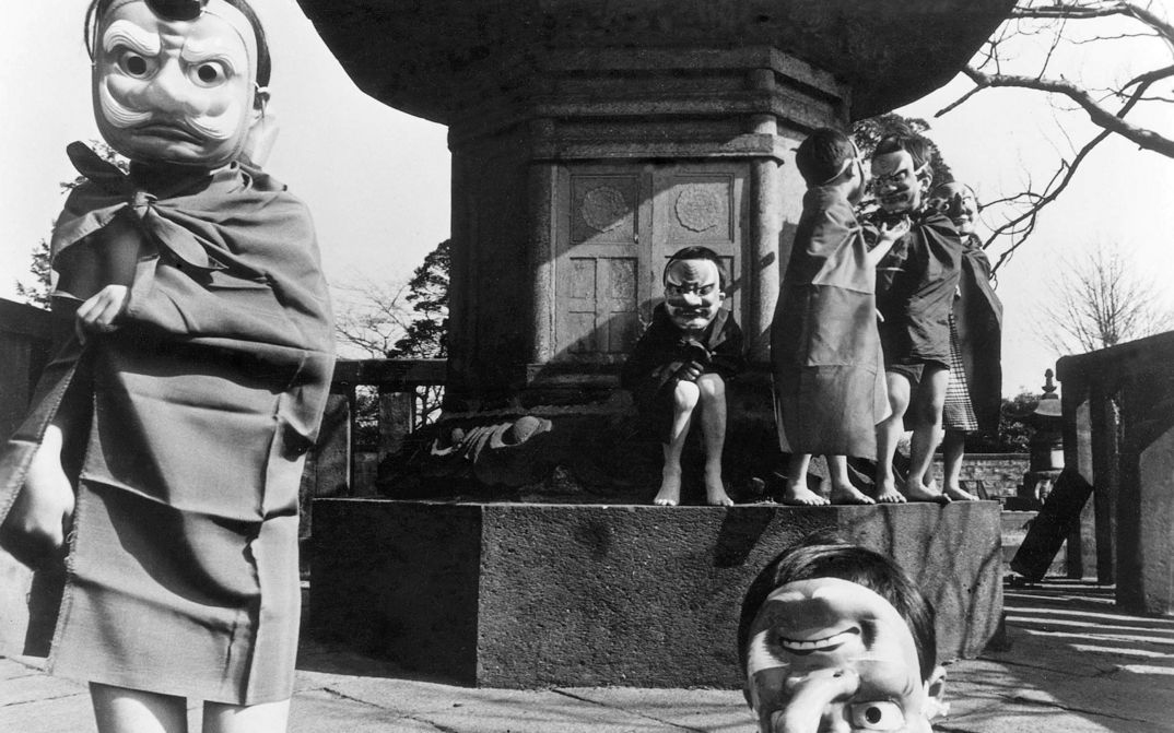 Film still from NANAMI, THE INFERNO OF FIRST LOVE: A group of children wearing masks stand and sit in front of a monument.