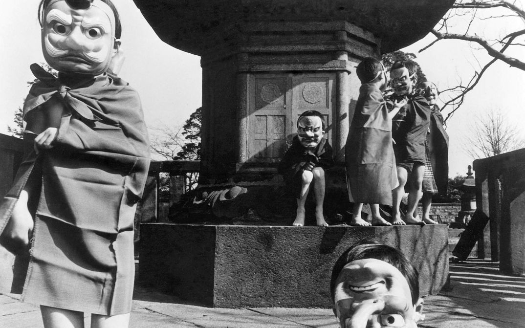 Film still from NANAMI, THE INFERNO OF FIRST LOVE: A group of children wearing masks stand and sit in front of a monument.