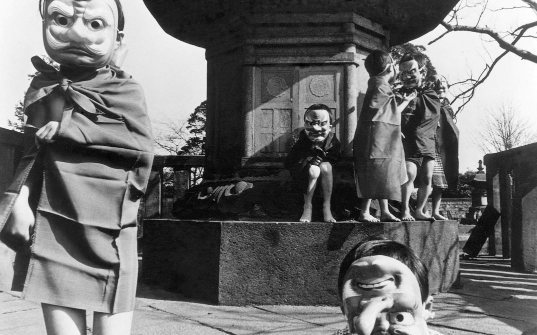 Filmstill aus DAS MÄDCHEN NANAMI: Eine Gruppe von Kindern mit Masken stehen und sitzen vor einem Denkmal.