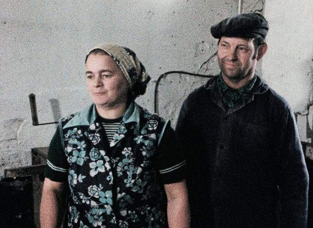 Still from the film "Ein Herbst im Ländchen Bärwalde" by Gautam Bora. A women with a patterned headscarf and a vest with blue flowers on it is standing in front of a white wall next to a man in a dark cap and jacket.