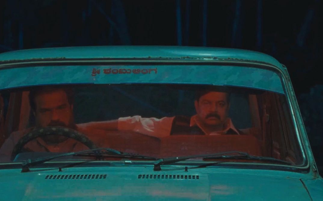 Film still from VAGHACHIPANI by Natesh Hegde. A view through the windshield shows two men in a car.